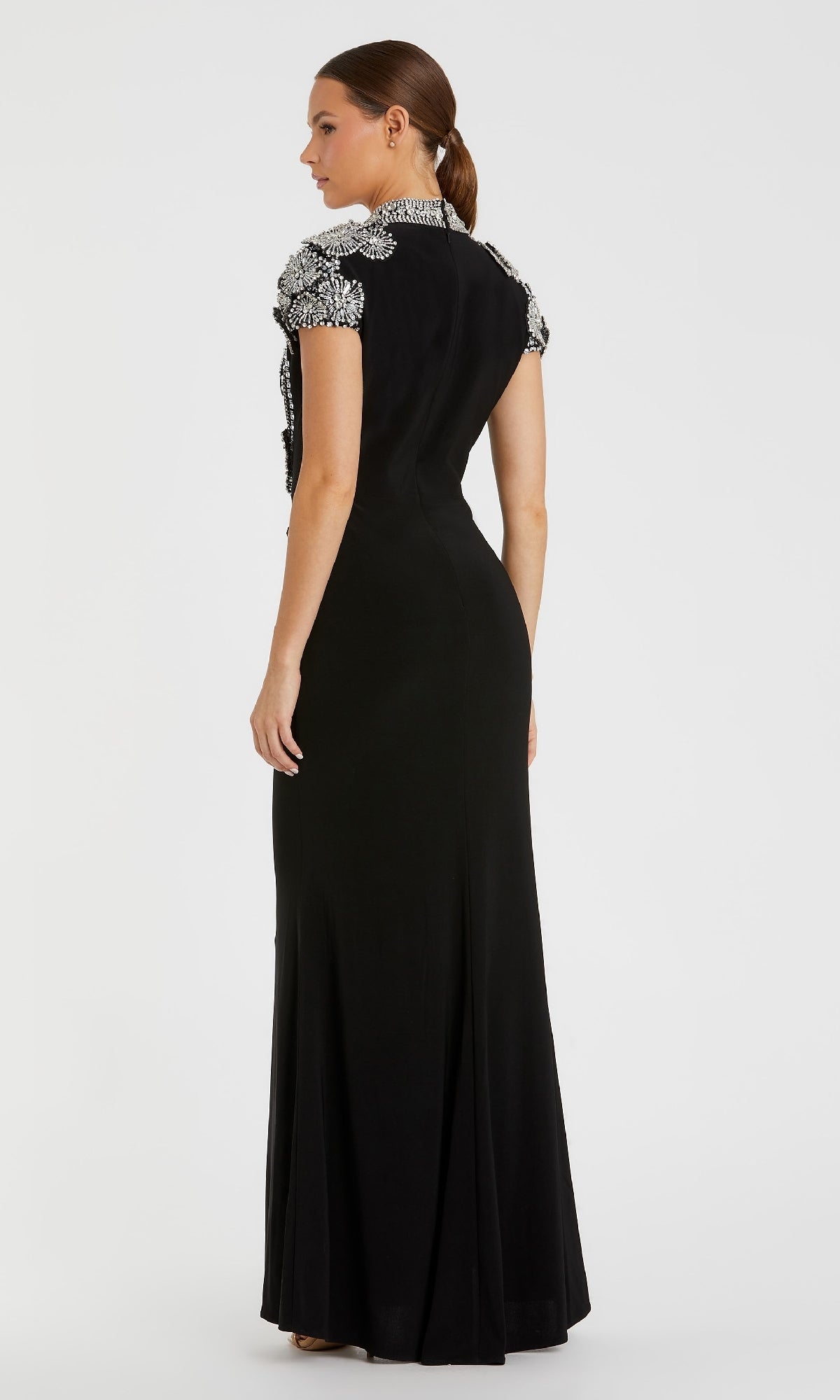 A woman stands facing away in the Mac Duggal 42055 beaded long black formal dress, featuring silver embellishments on the shoulders and collar, set against a plain white background.
