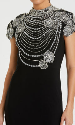 A woman wears the Mac Duggal 42055 beaded long black formal dress, featuring intricate silver bead and crystal floral embellishments with draped patterns on the bodice and shoulders.