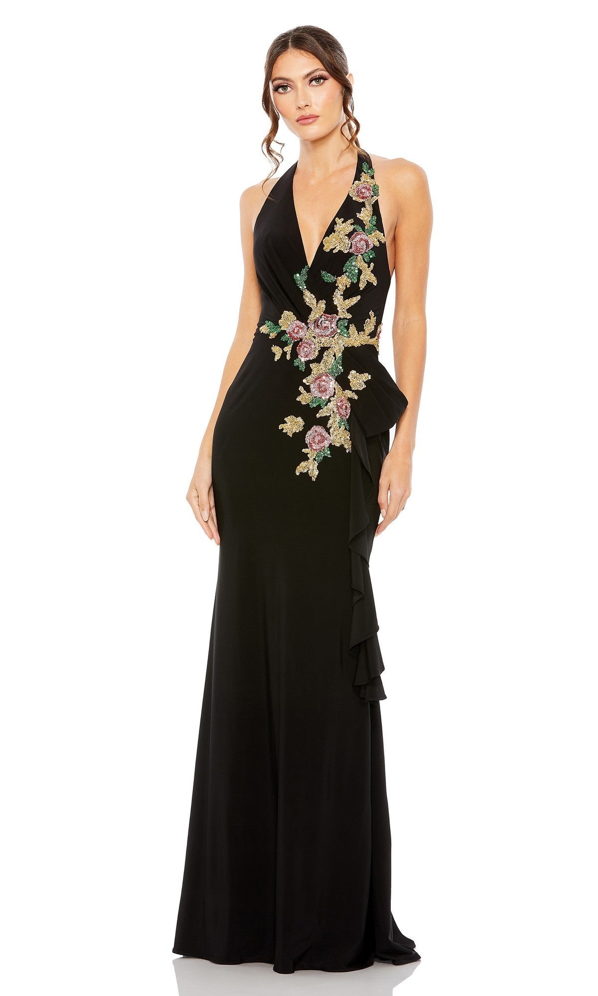 A woman models the Mac Duggal 42056 long formal dress in black, featuring a halter neckline, intricate floral appliques on the bodice and waist, and ruffle detailing along the side, as she poses against a white background.