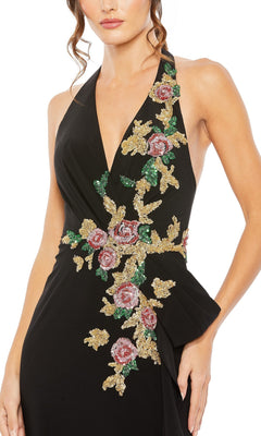 A woman wearing the Mac Duggal 42056 long formal dress, a sleeveless black halter gown with a deep V neckline, adorned with pink and gold floral appliques on the front—showcasing signature Mac Duggal elegance.