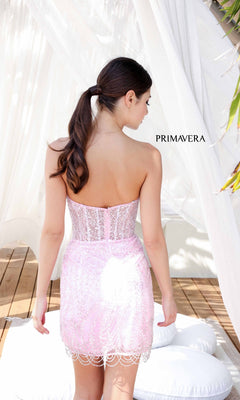 A woman with brown hair in a ponytail wears the Short Homecoming Dress 4213 by Primavera in light pink lace, standing with her back to the camera near white curtains and cushions. "PRIMAVERA" is visible in the background.