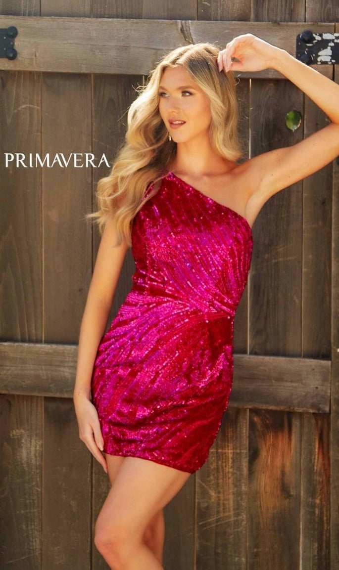 A woman poses outdoors in front of a wooden fence wearing the Primavera 4214 One-Shoulder Beaded Hoco Dress. The word 