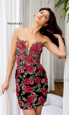 A woman poses indoors near white curtains wearing the Short Homecoming Dress 4215 by Primavera—a black strapless mini dress with red and pink floral embroidery. "PRIMAVERA" appears on the left, highlighting the rose-embellished design.