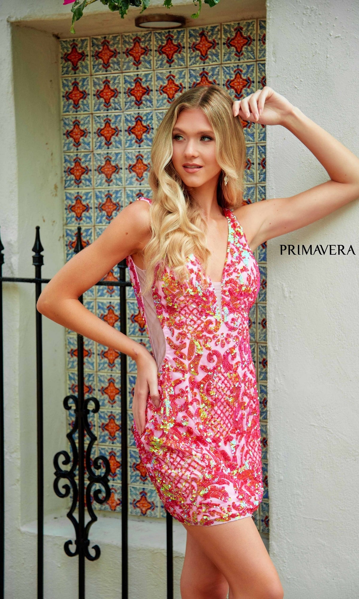 A woman wearing the Backless Short Sequin Hoco Dress: Primavera 4218, featuring pink and orange patterns, stands before a decorative tile wall with 