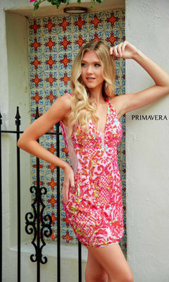 A woman wearing the Backless Short Sequin Hoco Dress: Primavera 4218, featuring pink and orange patterns, stands before a decorative tile wall with "PRIMAVERA" displayed beside her and a black fence in the background.
