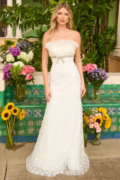 A woman wearing the Ava Presley 42224 long prom dress stands outdoors in front of colorful floral arrangements and green tiled walls.