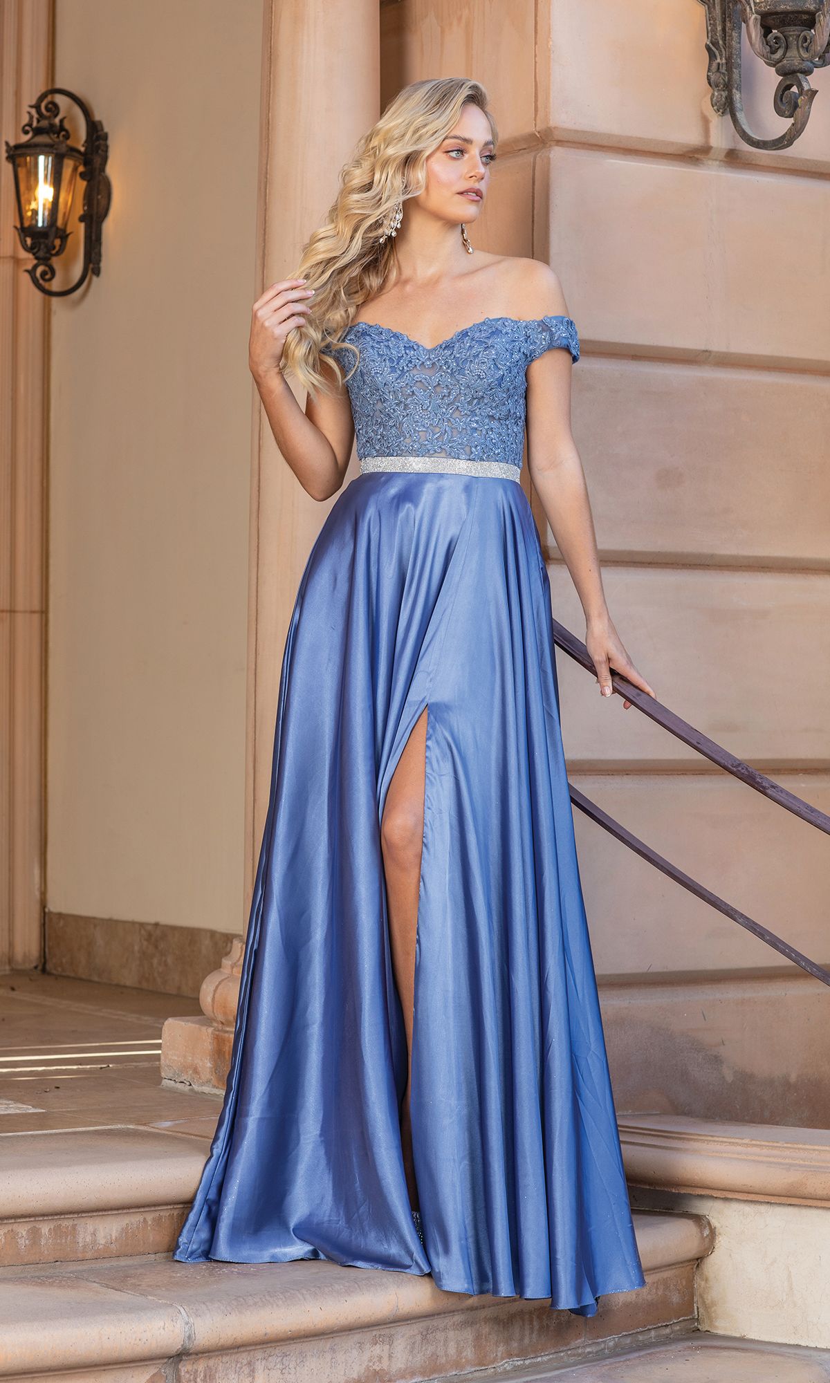 A woman wearing the Embroidered Off-Shoulder Long Prom Dress 4222A, with a sheer embroidered bodice and thigh-high slit, stands on steps near a handrail and ornate wall sconce.