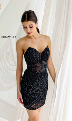 A woman stands by white curtains wearing the Short Homecoming Dress 4222 by Primavera—a strapless black mini dress featuring sequined beading, sheer details, and a corset bodice. "PRIMAVERA" appears on the left side.