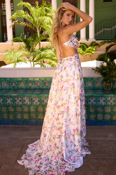 A woman in an open back stands outdoors by a tiled wall, wearing the Long Prom Dress: Ava Presley 42236—a stunning floral chiffon gown perfect for elegant occasions.