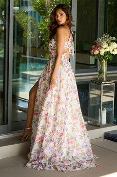 A woman in the Ava Presley 42237 long prom dress, featuring floral chiffon and a high slit, stands by a glass wall and looks over her shoulder with flowers and greenery in the background.