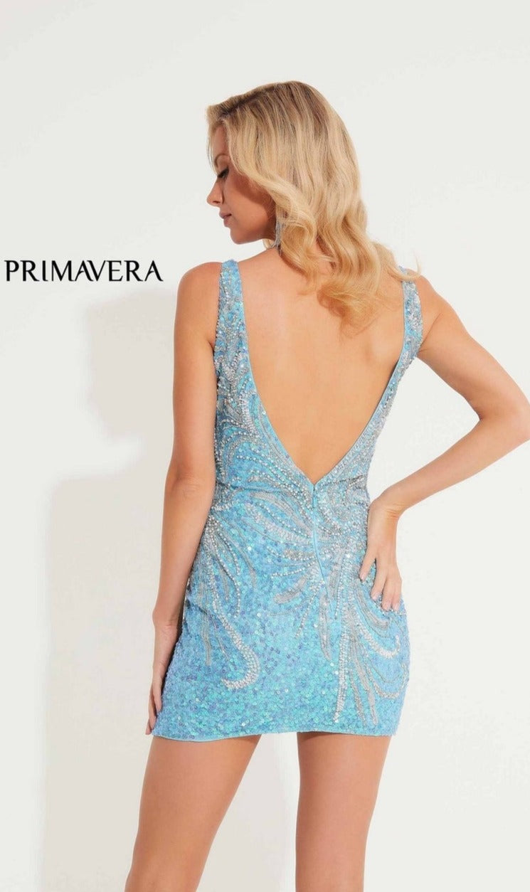A woman with wavy blonde hair models the Tulip-Hem Beaded Mini Hoco Dress: Primavera 4223—a blue, backless party dress with sequins and a deep V-back—against a plain white background. 