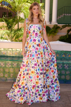 A woman stands outdoors by a tiled wall and greenery, wearing the Ava Presley 42266: a long, sleeveless white prom dress with a colorful floral print.