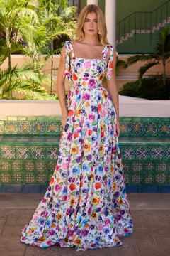 A woman stands outdoors in the Ava Presley 42266 long floral-print prom dress, surrounded by lush green plants and patterned tiles for a charming scene.
