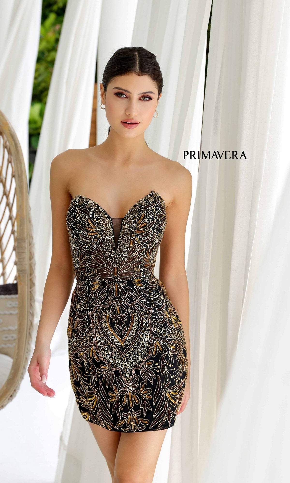 A woman stands indoors near white drapes wearing the Strapless Short Beaded Hoco Dress: Primavera 4226—a fitted black mini with gold and silver embroidery.