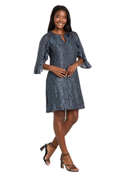 A woman smiles in the R&M Richards 4226 Short Lace Semi-Formal Dress, featuring a knee-length navy a-line silhouette and three-quarter sleeves, paired with black heeled sandals against a white background.