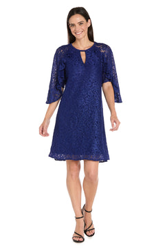 Woman models the R&M Richards 4226 Short Lace Semi-Formal Dress, featuring knee-length blue glitter lace, half sleeves, and paired with black strappy heels, smiling against a plain white background.