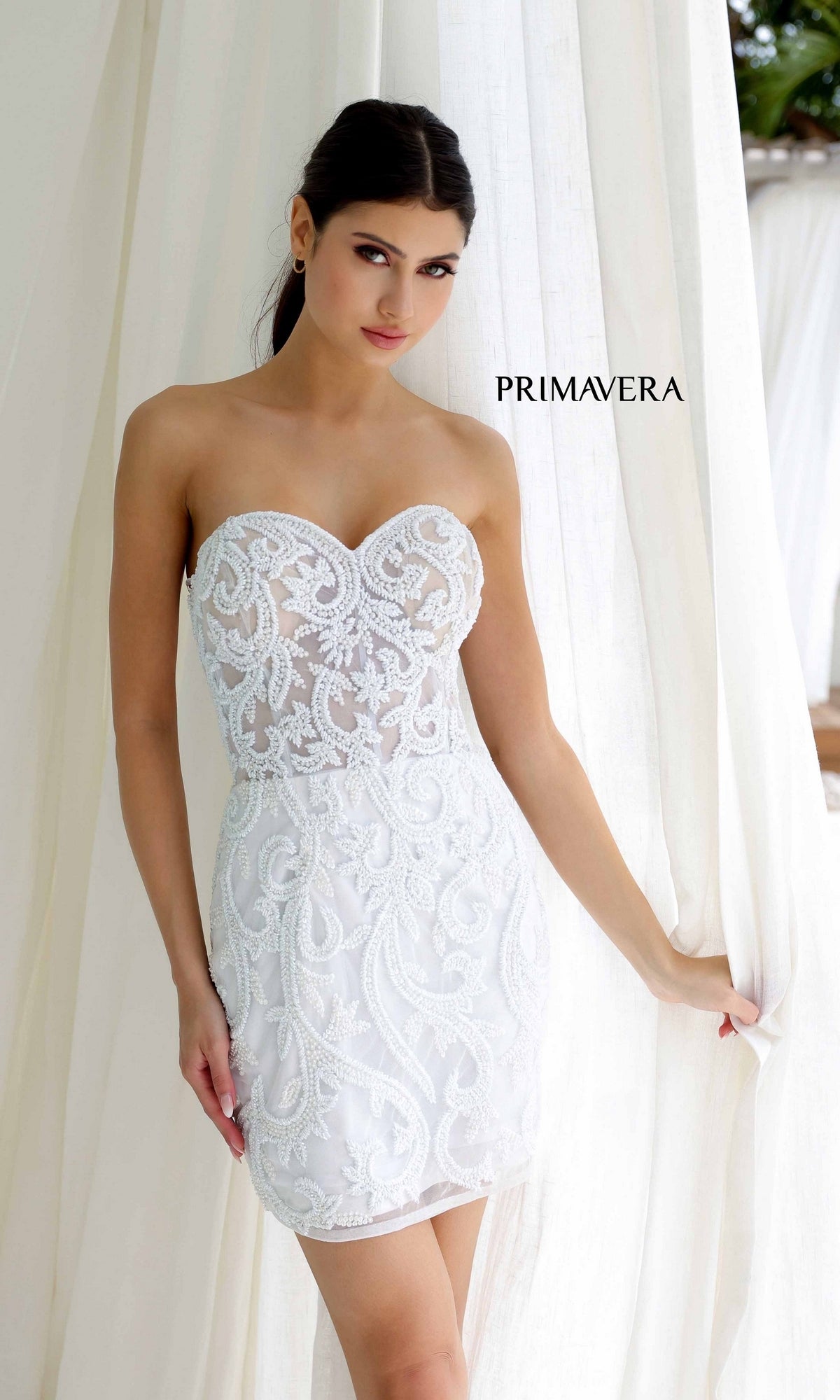 A woman in a white, strapless beaded short homecoming dress, Primavera 4227, stands by white curtains with 