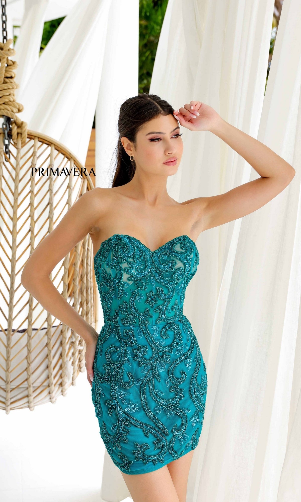 A woman in the Strapless Beaded Short Hoco Dress Primavera 4227 poses with one arm raised, standing by white curtains and a hanging chair.
