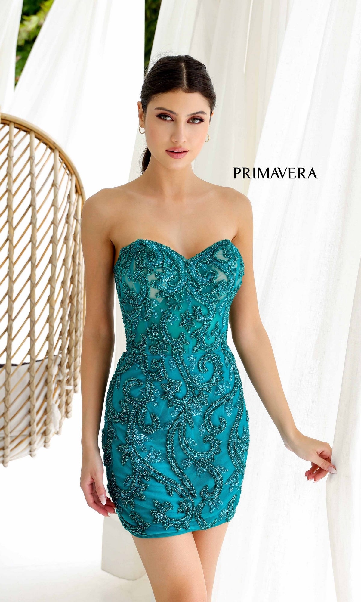 A woman models the Strapless Beaded Short Hoco Dress: Primavera 4227 in teal, with intricate beadwork, standing indoors near white curtains and a wicker chair. 