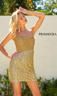 A woman in the Strapless Fringe Hoco Dress: Primavera 4229, featuring a beaded short design with a gold sequin-print bodice, poses outdoors with her hand near her head. "PRIMAVERA" is visible on the wall behind her.