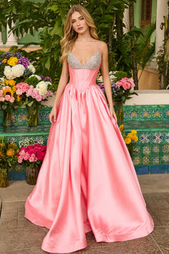 Wearing the Ava Presley 42311 long prom dress in pink satin with a jeweled corset bodice, a woman stands outdoors by colorful flower arrangements and decorative tiles.