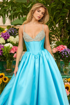 A woman stands before vibrant flowers wearing the Ava Presley 42311 long prom dress—a shiny light blue ball gown with a fitted, jeweled bodice and thin straps.