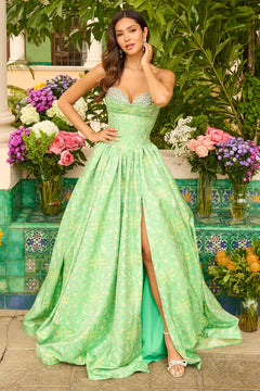 A woman stands outdoors in the green Ava Presley 42318 long prom dress, a strapless gown with a high slit and floral print, surrounded by colorful flowers and lush greenery.