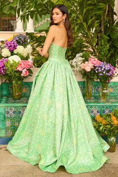A woman wearing the Ava Presley 42318 long prom dress with a strapless corset bodice stands outdoors among lush greenery and vibrant flower arrangements.