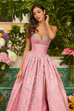 A woman wearing the Ava Presley 42318 long prom dress with a beaded bodice stands outdoors amid lush greenery and vibrant flowers.