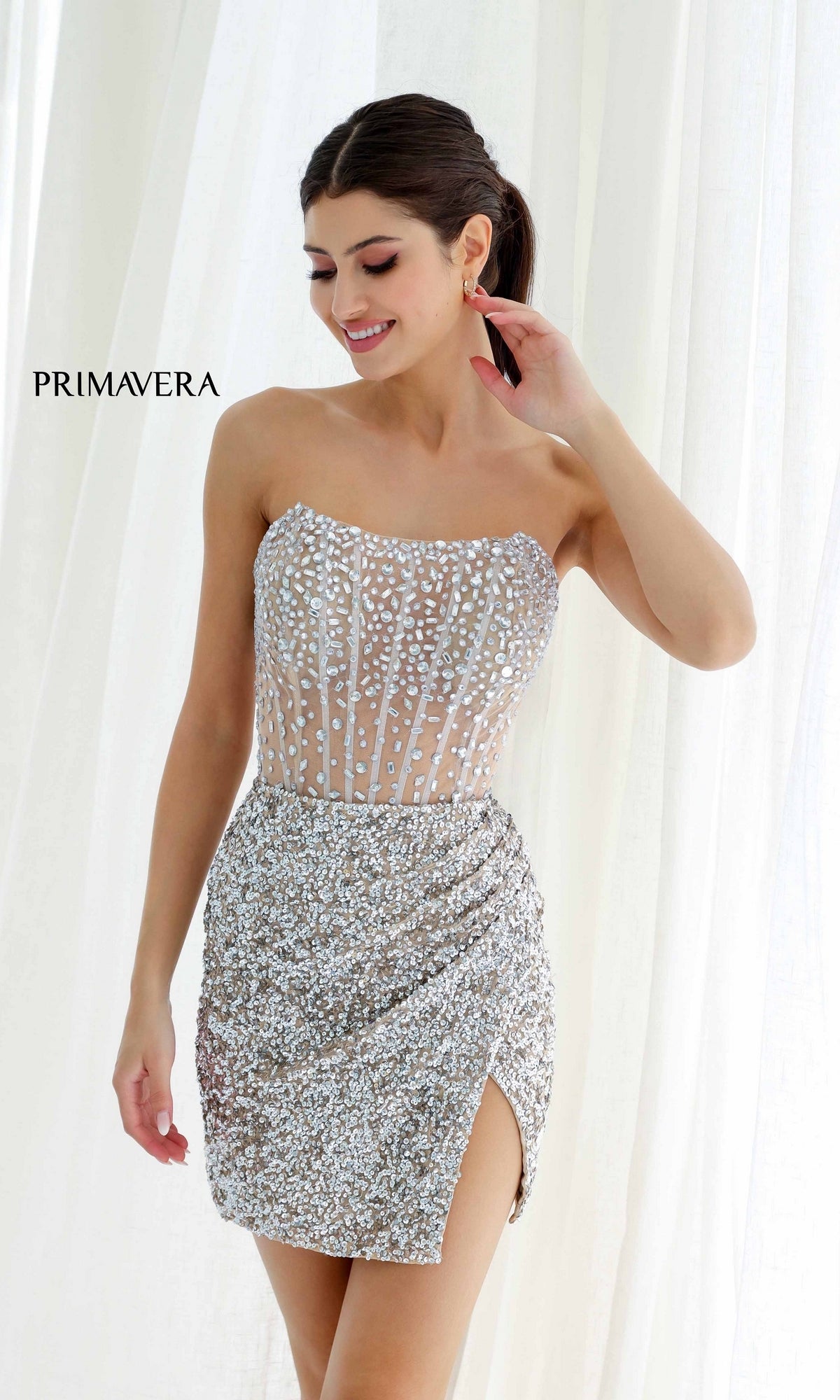 A woman wears the Short Homecoming Dress 4231 by Primavera, a strapless fitted mini party dress with silver sequins, a beaded slit, and sheer boned corset detail, posed before white curtains with 