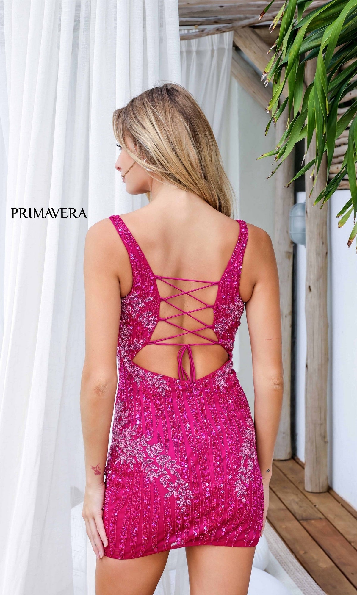 A woman stands with her back to the camera, showcasing the Short Homecoming Dress 4232 by Primavera—a pink sequined dress with lace-up back—set amid white curtains and a wooden structure.