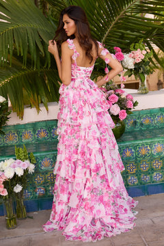 A woman stands outdoors before palm leaves, wearing the elegant Ava Presley 42332 long prom dress with floral chiffon, a low back, and ruffled details. Pink and white flower vases nearby add romance to the scene.