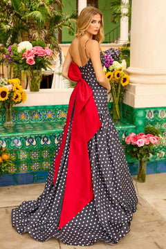 A woman wearing the Ava Presley 42368 Long Prom Dress—a black and white polka dot gown with a large red bow—stands by colorful flowers and green tiled columns outdoors.