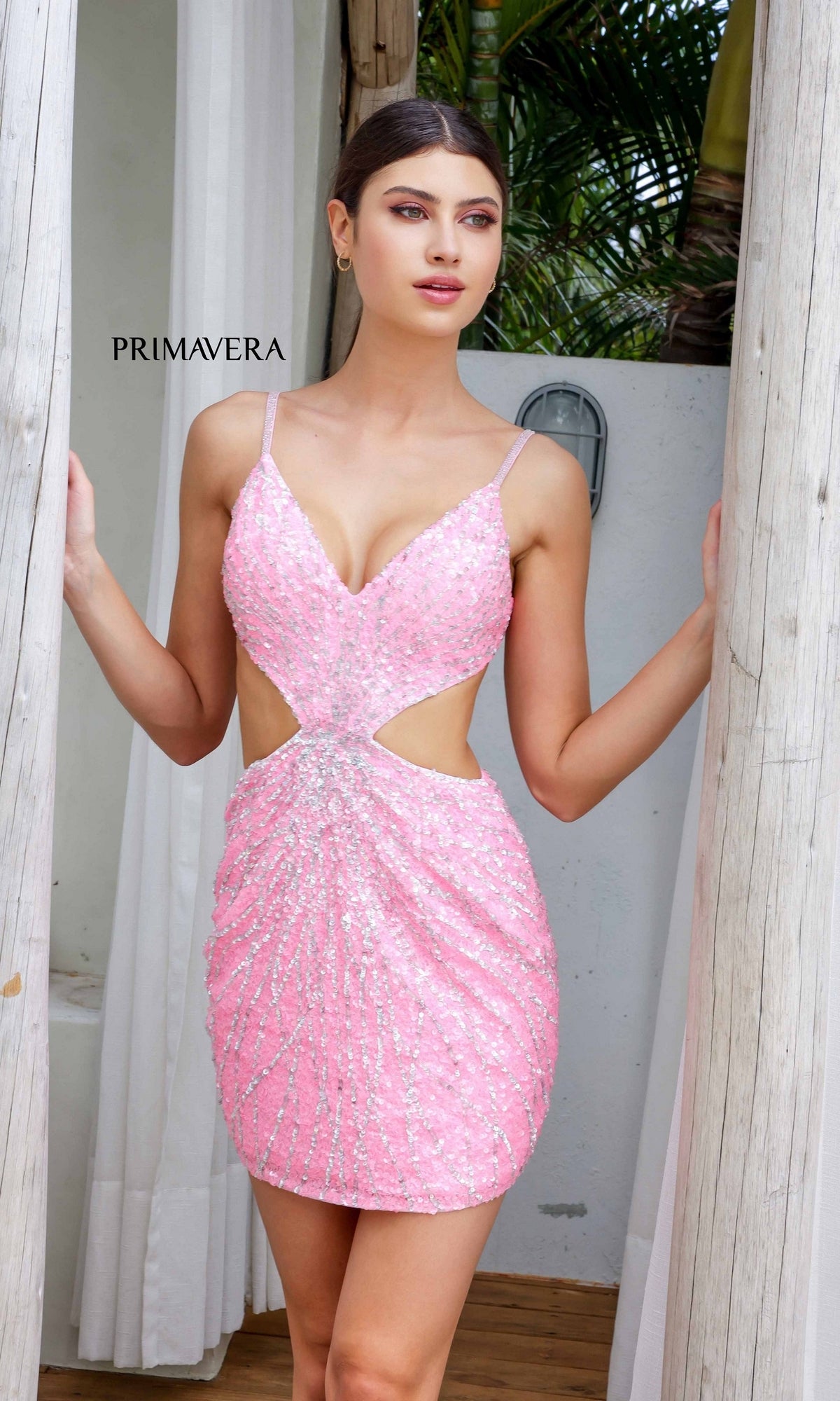 A woman stands between wooden posts, looking to the side, wearing the Short Homecoming Dress 4236 by Primavera—a pink sequin mini dress with cut-out sides and an open back. White walls, curtains, and lush tropical plants are in the background.