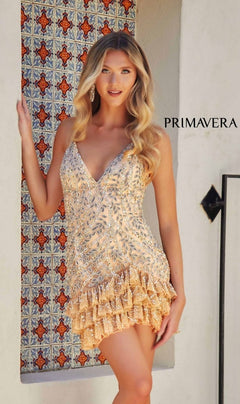 A woman stands before a patterned tile backdrop, wearing the Short Homecoming Dress 4242 by Primavera—a gold sequined mini dress with a lace-up back and ruffled hem. "PRIMAVERA" is displayed on the wall behind her.