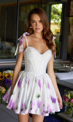 A woman stands indoors wearing the Ava Presley 42432 short cocktail dress, featuring a floral print satin fabric, pearl-embellished corset, and ribbon-tied shoulder straps. Flowers and greenery decorate the background.