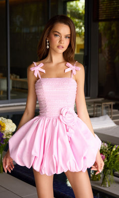 A woman stands indoors wearing the Ava Presley 42449 short homecoming dress in light pink, featuring a fit-and-flare silhouette, bow straps, pearl embellishments, and a large flower at the waist.