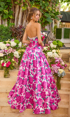 A person stands on stone steps wearing an Ava Presley 42459 floral, strapless, floor-length ball gown, surrounded by large bouquets of flowers.