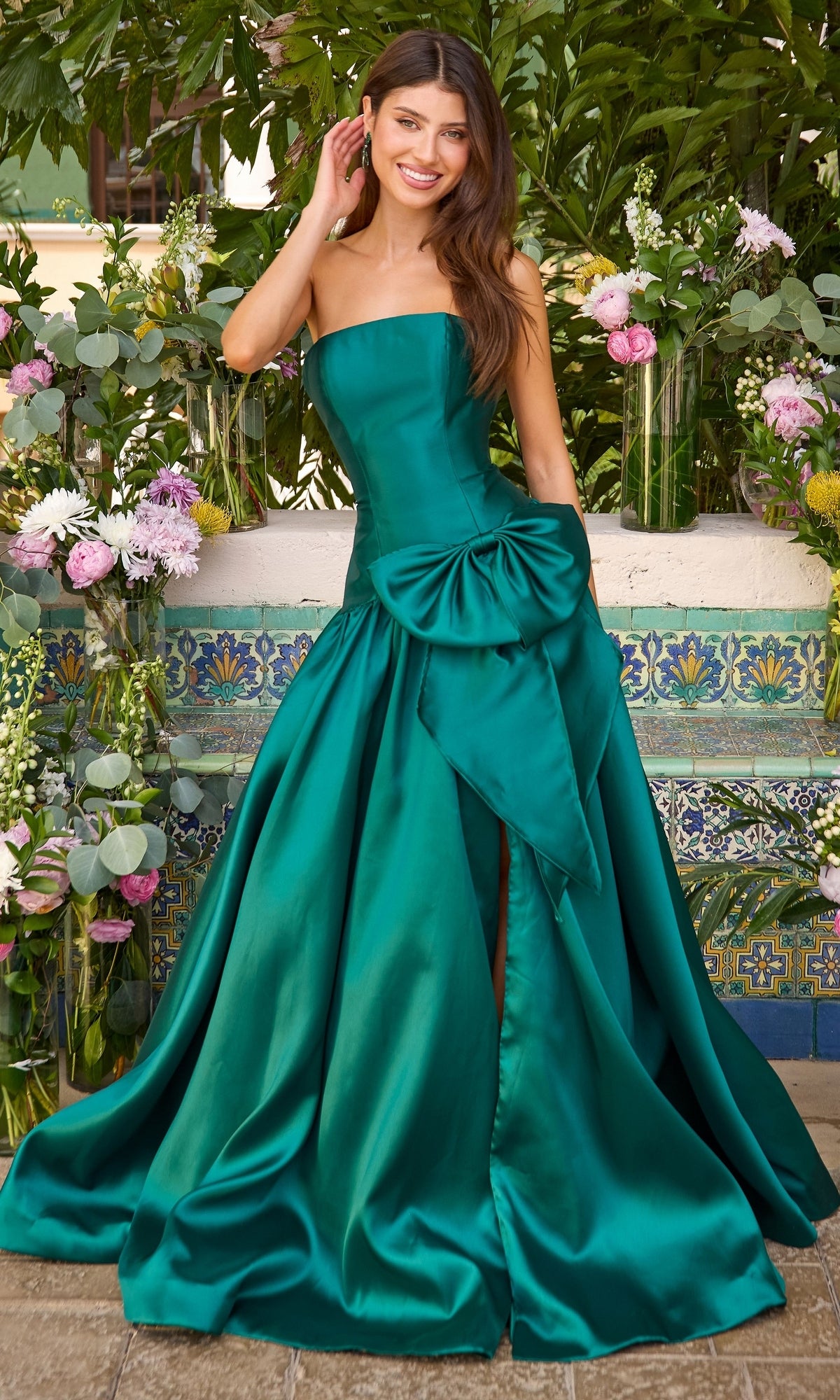A smiling woman wears an Ava Presley 42477 hunter green strapless prom ball gown featuring a large bow at the waist and a thigh-high slit.