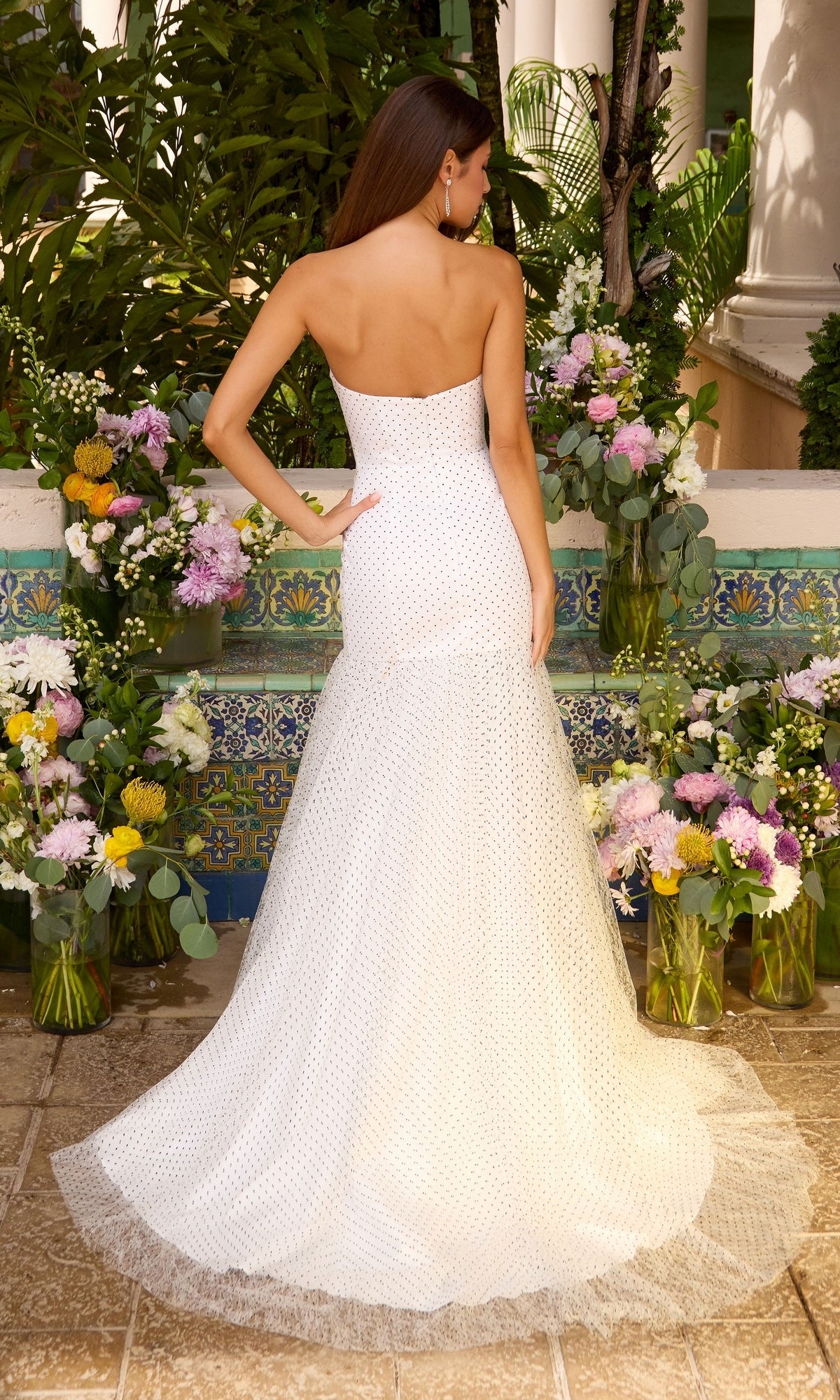 A woman seen from behind wearing an Ava Presley 42489 strapless, floor-length white polka dot prom dress with a small train, standing in front of floral arrangements.