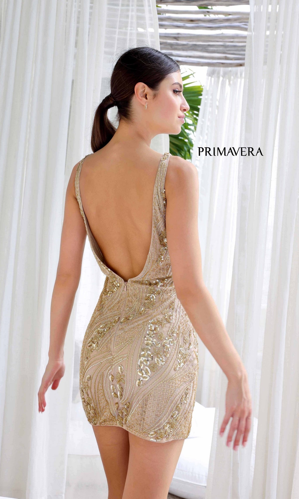 A woman wears the Short Homecoming Dress 4249 by Primavera—a gold, beaded, open-back mini dress—while standing indoors near sheer white curtains. 