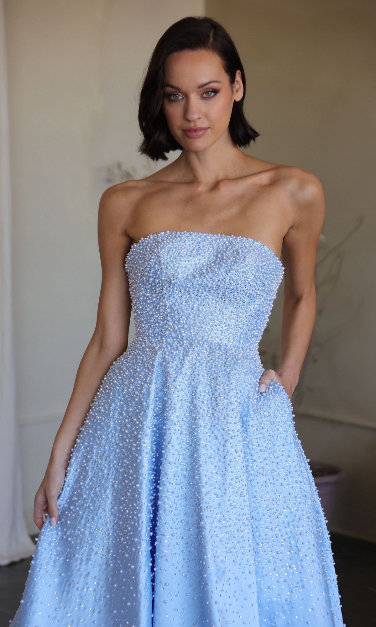A woman models the Ava Presley 42500 long prom dress in light blue, featuring a strapless design with small white beads, standing indoors against a neutral background.