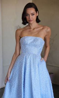 A woman models the Ava Presley 42500 long prom dress in light blue, featuring a strapless design with small white beads, standing indoors against a neutral background.