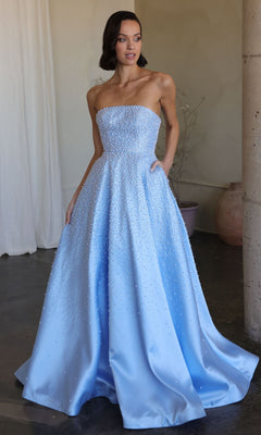 A woman stands indoors wearing the Ava Presley 42500, a long prom dress featuring a strapless, fitted bodice, floor-length light blue skirt, and subtle beaded embellishments.