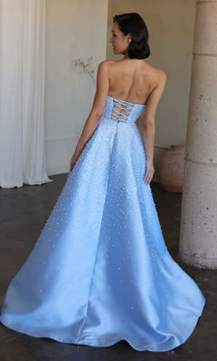 A woman in the Ava Presley 42500 long prom dress—a light blue, strapless, floor-length gown with a lace-up back and beaded details—stands facing away in an elegant indoor setting.