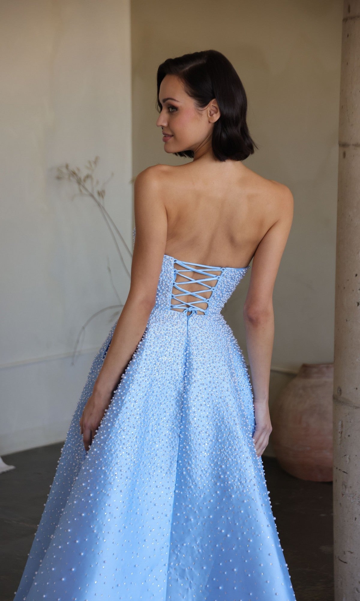 A woman in the Ava Presley 42500 long prom dress, featuring a strapless light blue design with a lace-up back and pearl-like embellishments, stands indoors facing slightly away from the camera.