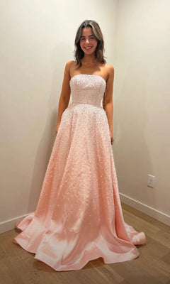 A young woman stands in a simple room wearing the Ava Presley 42500, a stunning blush, strapless long prom dress adorned with beautiful pearls.
