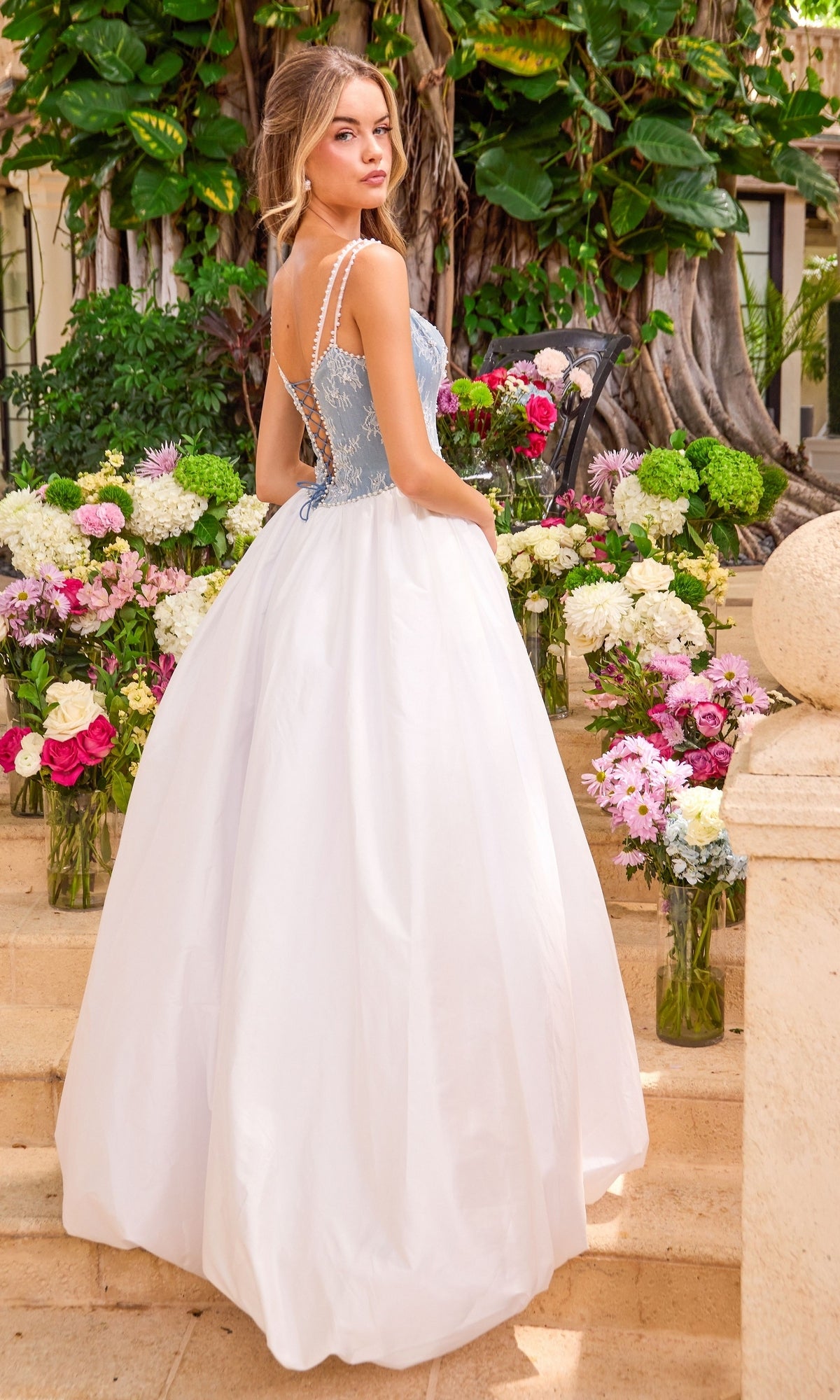 The back view of a blue and white Long Prom Dress: Ava Presley 42519 featuring a lace corset bodice and full skirt, positioned on stone steps in front of flower arrangements.