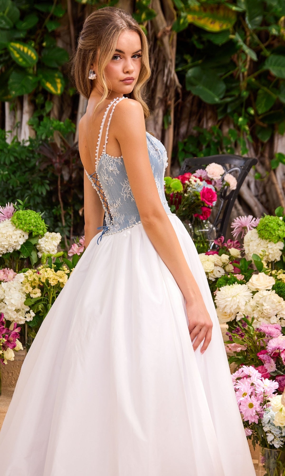 A woman wears an Ava Presley 42519 long prom dress featuring a blue lace corset-style bodice with pearl-beaded straps and a flowing white skirt.