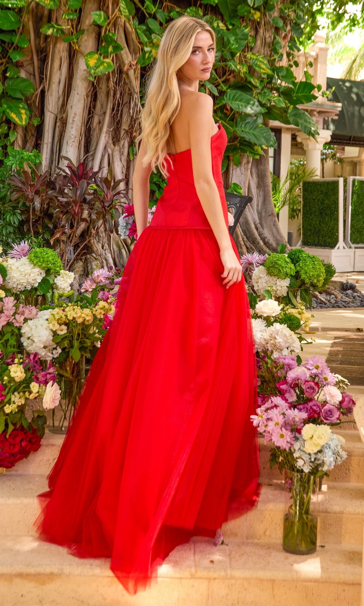 A woman in a red strapless Ava Presley 42521 A-line gown stands on stone steps surrounded by vases of flowers.